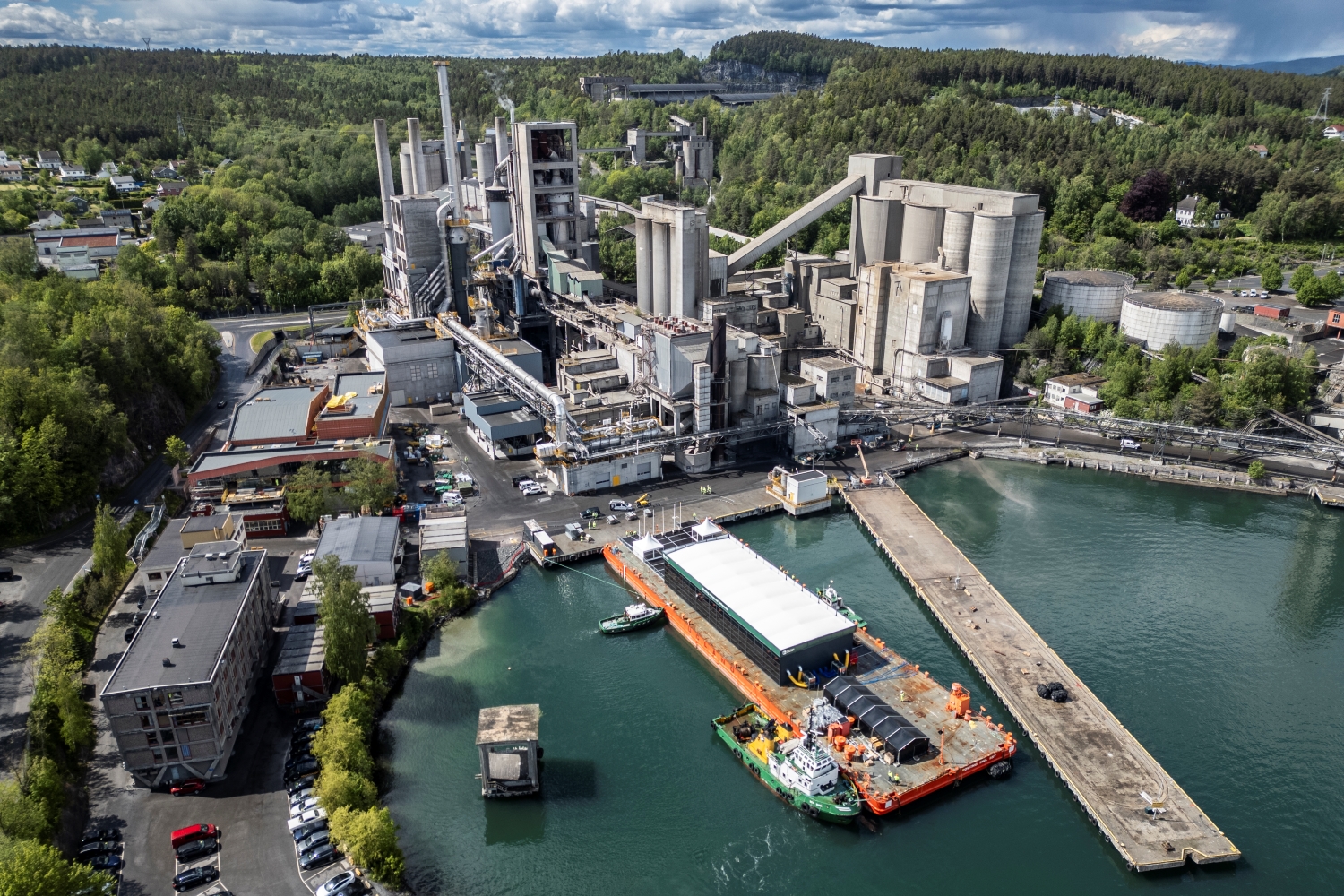 Aerial view of a cement plant complex near a waterfront, surrounded by green forested hills. The facility includes large silos, conveyor belts, and warehouse buildings. A docked barge with containers is visible in the water alongside a long pier, with small boats nearby. Residential and office buildings are located to the left of the factory.