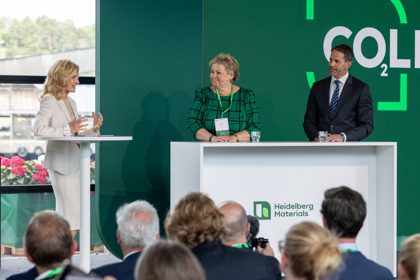 Kvinne i hvit drakt står til venstre og taler til to personer bak et podium, inkludert Erna Solberg (midt) i grønn rutete kjole. På scenen vises logoen til Heidelberg Materials og teksten "CO2LL". Publikum sitter foran i bildet. Scenen er innendørs med et vindu og grønn natur utenfor.