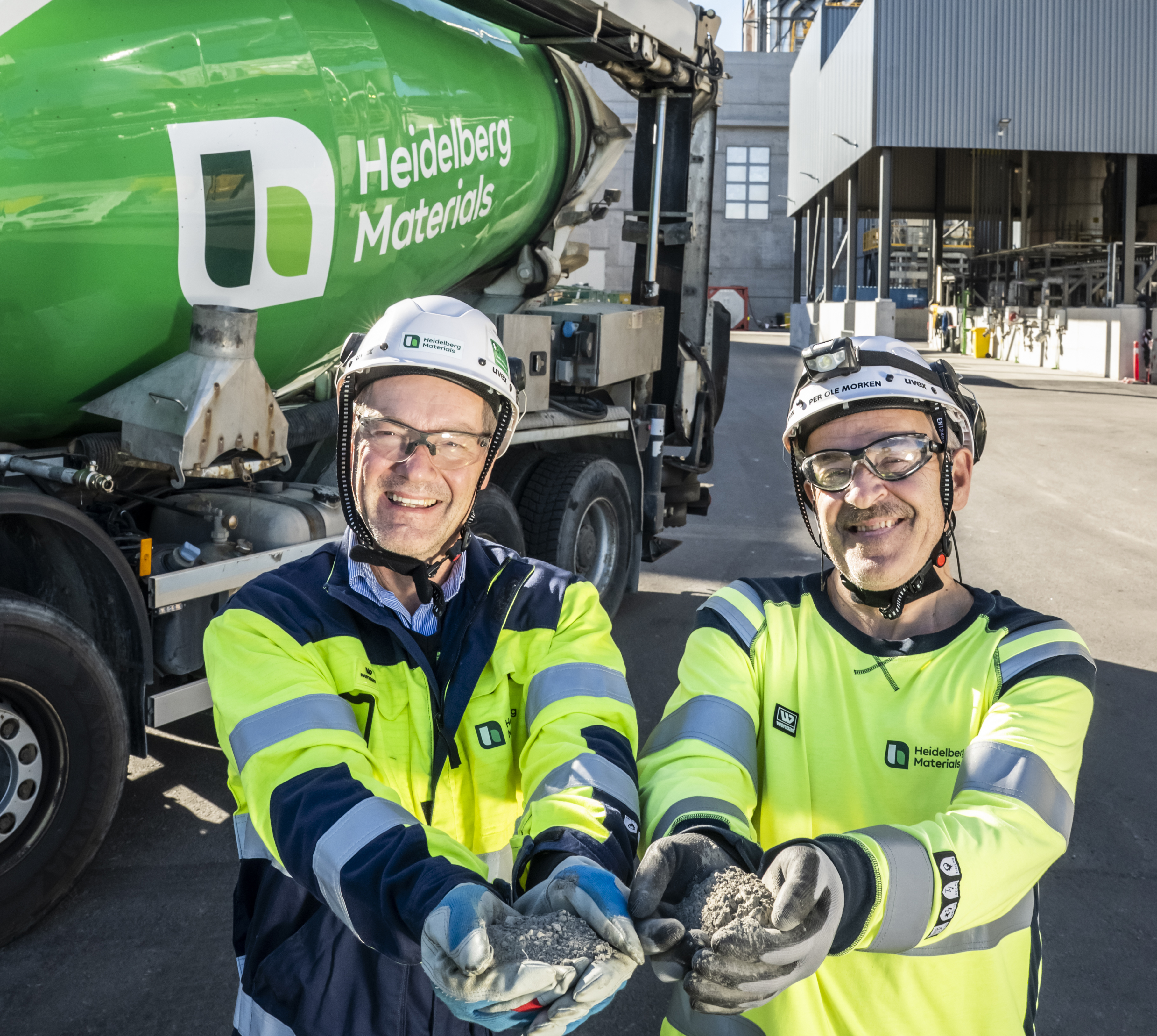 To personer i arbeidsklær og verneutstyr står foran en grønn betongbil med logoen "Heidelberg Materials". De holder frem to never med grå betongmateriale. Fabrikkbygninger og arbeidsområde vises i bakgrunnen.